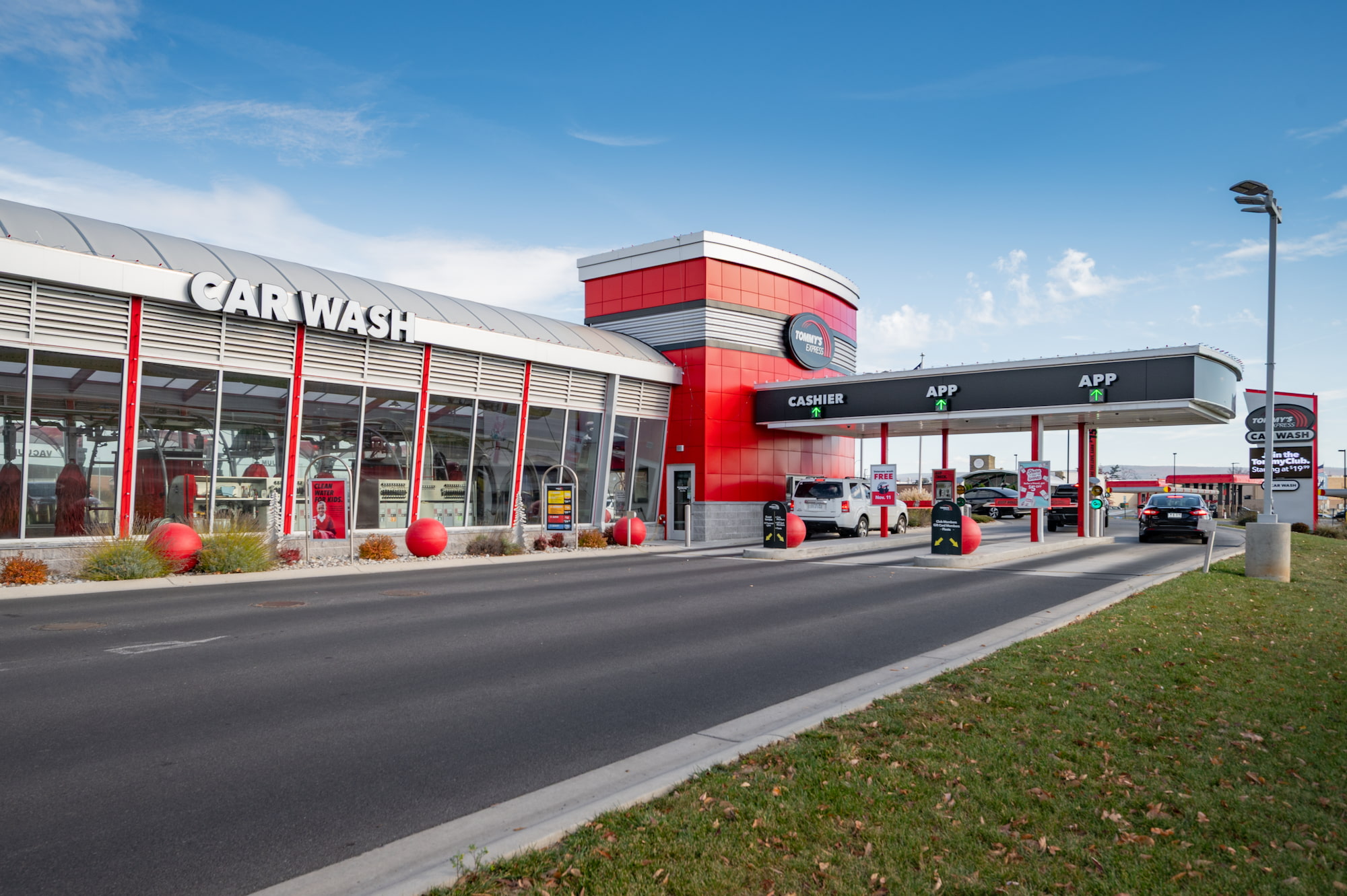 The Chambersburg Tommy’s Express Car Wash from the street, showing the glass-walled wash tunnel, the cashier and app pay lanes under the canopy, and cars queuing to enter
